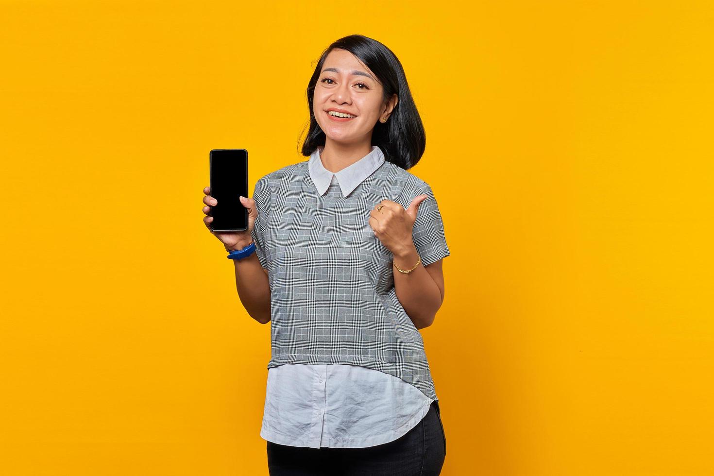 femme asiatique souriante montrant un écran de smartphone vierge et un pouce levé sur fond jaune photo