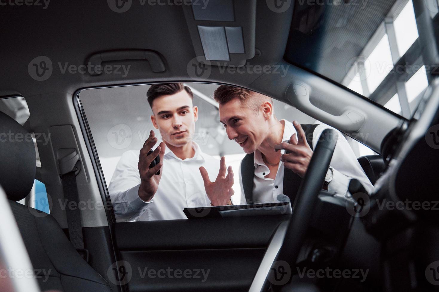 deux hommes se tiennent dans la salle d'exposition contre des voitures. gros plan d'un directeur des ventes en costume qui vend une voiture à un client. le vendeur remet la clé au client. photo