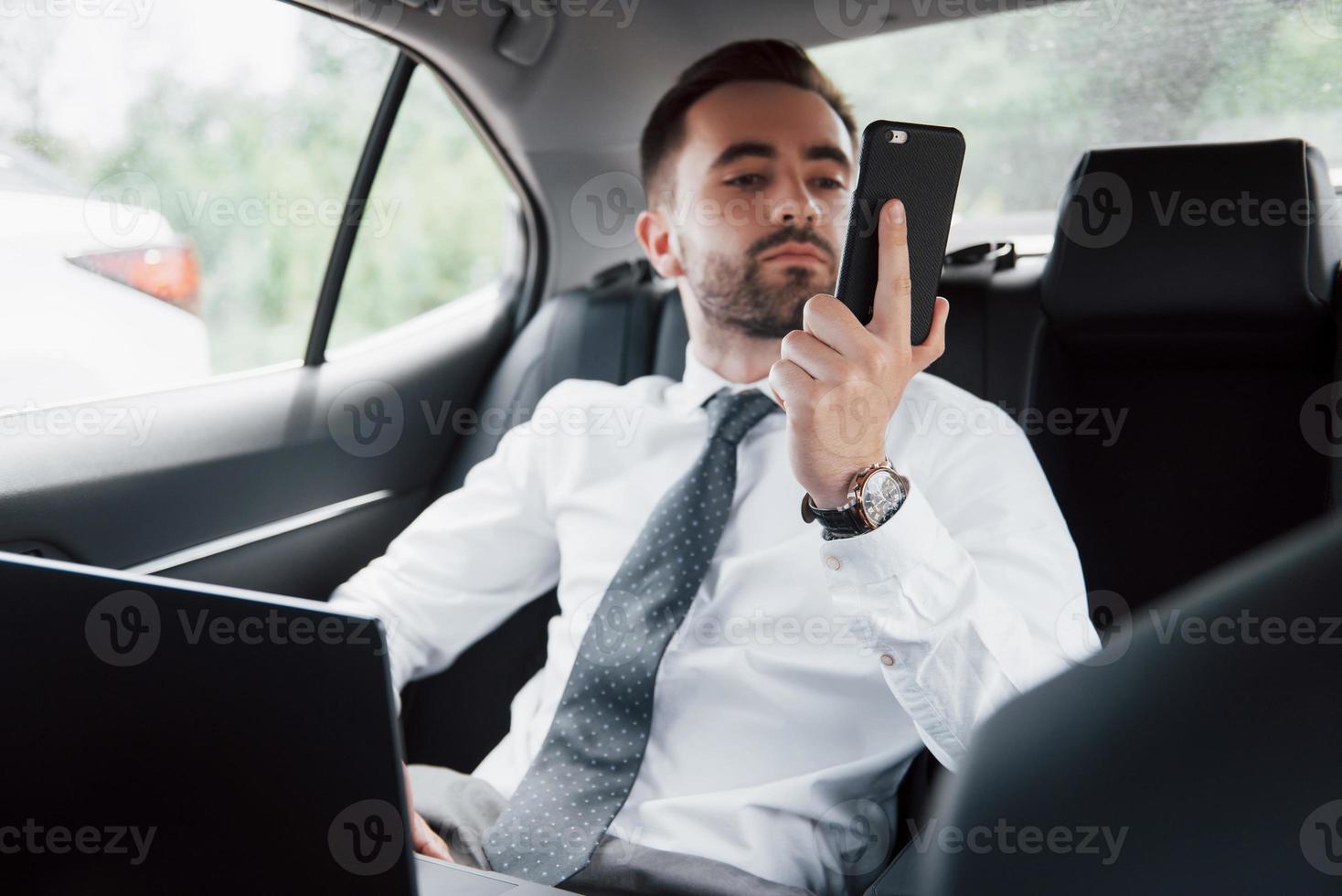 un jeune homme d'affaires travaillant sur un ordinateur portable et parlant au téléphone alors qu'il était assis à l'arrière de la voiture. travaille en mouvement, apprécie son temps photo