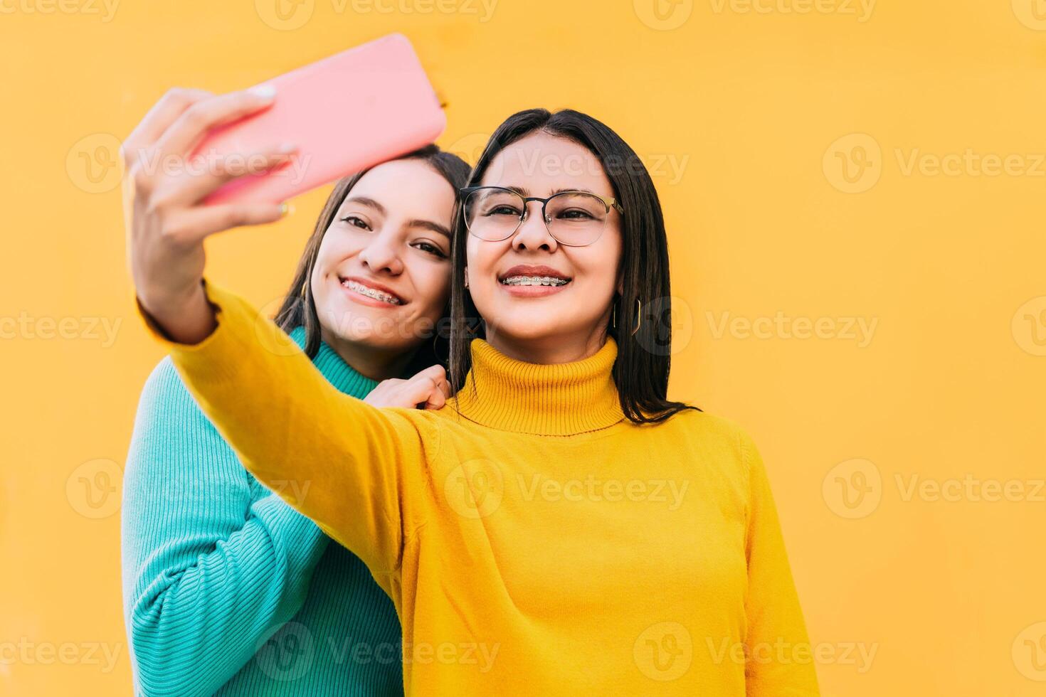 hispanique femmes dans bleu et Jaune chandails avec un appareil dentaire sur leur les dents prise une selfie. photo