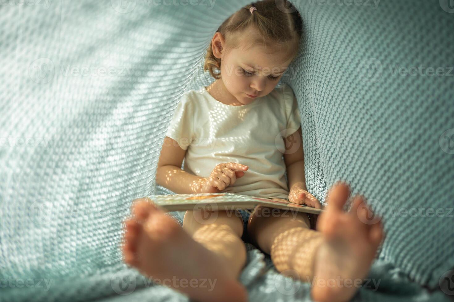 peu fille profiter de la revue une livre en dessous de bleu tricoté plaid dans ensoleillé Matin photo