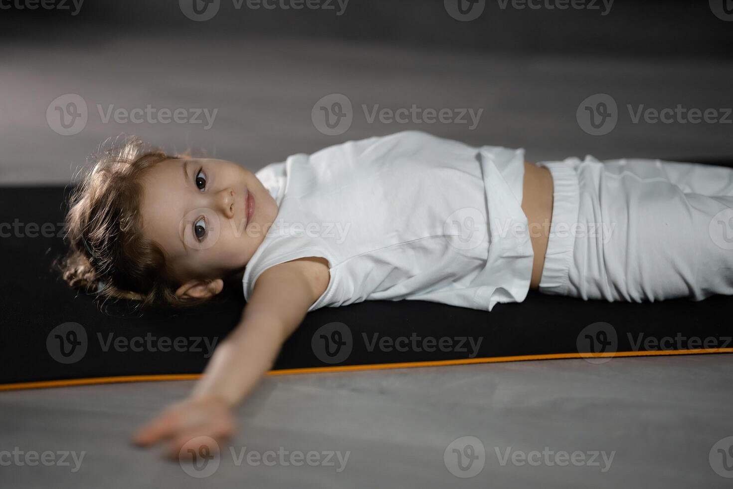 peu fille se détendre et méditer mensonge sur le tapis tandis que pratiquant yoga dans foncé pièce photo