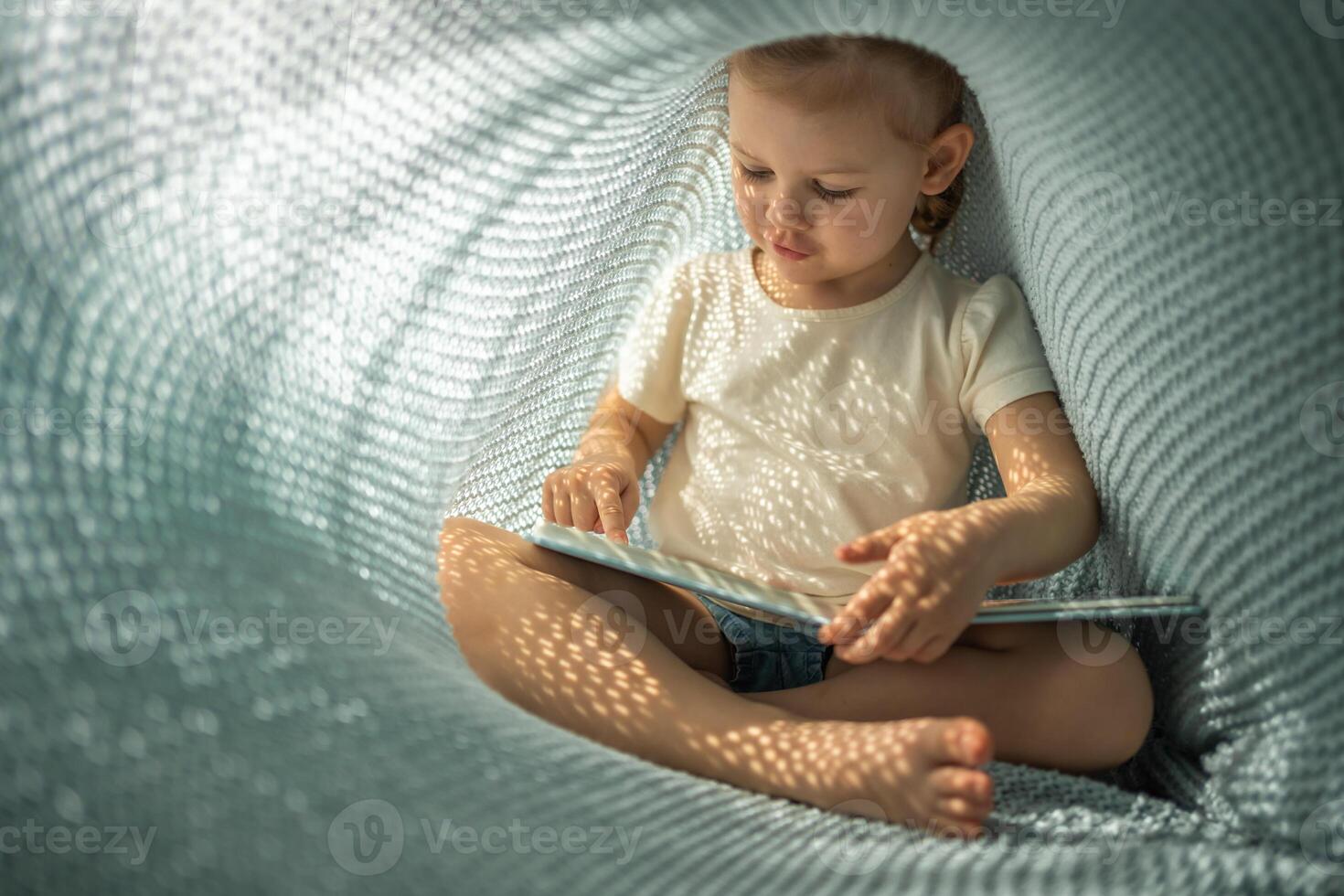 peu fille profiter de la revue une livre en dessous de bleu tricoté plaid dans ensoleillé Matin photo