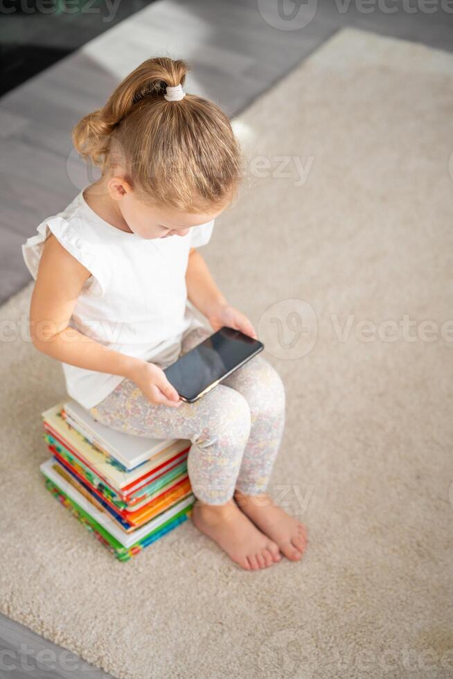 peu fille est assis sur une empiler de enfants Conte de fée livres et montres les dessins animés sur sa téléphone intelligent photo