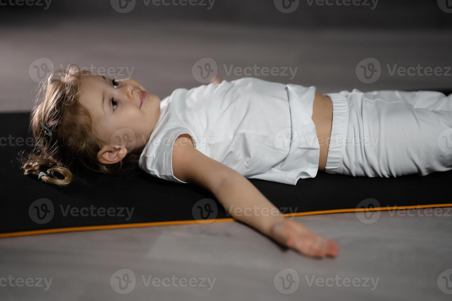 peu fille se détendre et méditer mensonge sur le tapis tandis que pratiquant yoga dans foncé chambre. haute qualité photo