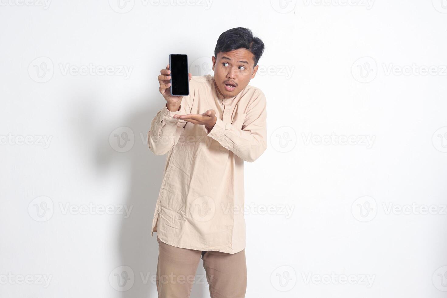 portrait de Jeune excité asiatique musulman homme dans koko chemise montrant Vide écran mobile téléphone maquette tandis que montrer du doigt et en présentant produit. social médias concept. isolé image sur blanc Contexte photo