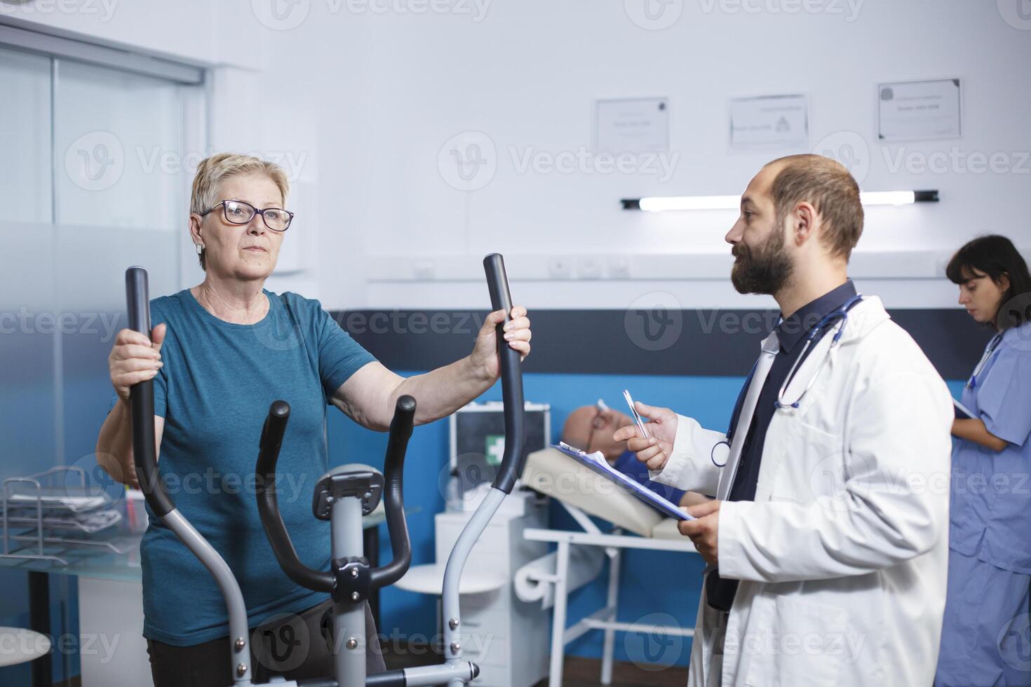 Masculin médecin parlant à Sénior patient Faire physique activité à récupérer de blessure avec électrique vélo. Sénior femme en utilisant Stationnaire vélo pour bien-être et récupération à physiothérapie clinique. photo