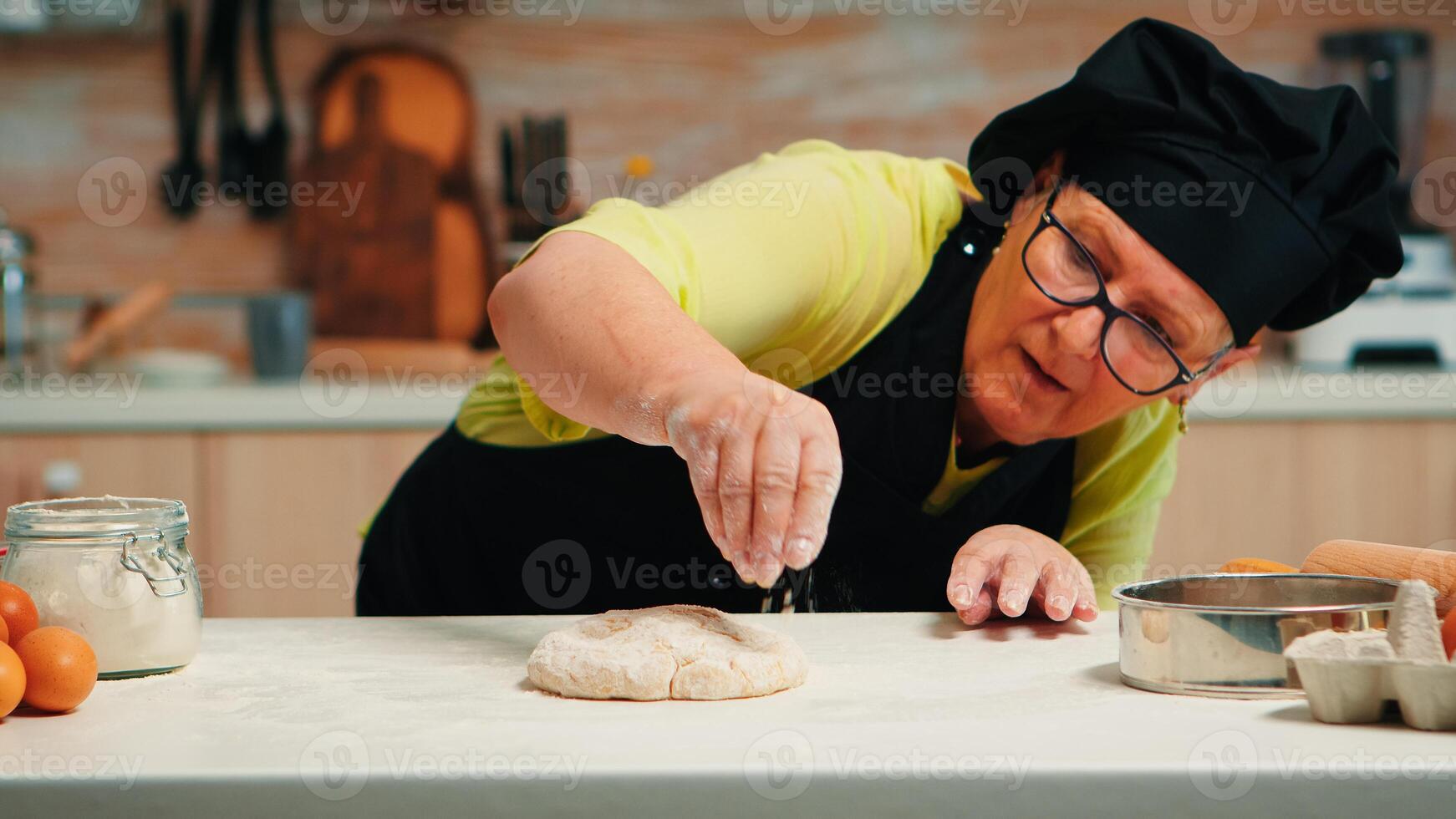 femme avec bonte et cuisine tablier est occupé avec pâte préparation. retraité Sénior boulanger avec tablier, cuisine uniforme saupoudrage, tamisage, diffusion farine avec main cuisson fait maison Pizza et pain. photo