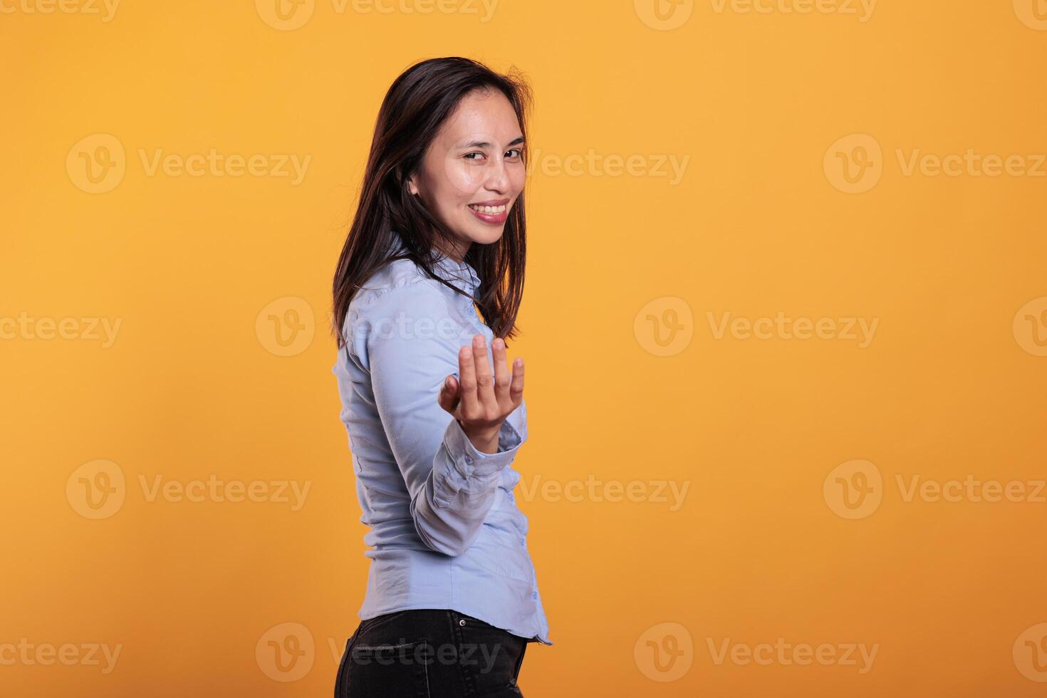 souriant de bonne humeur asiatique modèle Faire viens à moi symbole, faire signe avec doigt posant dans studio plus de Jaune Contexte. brunette femme attrayant vous pour confidentiel parler. appel geste photo