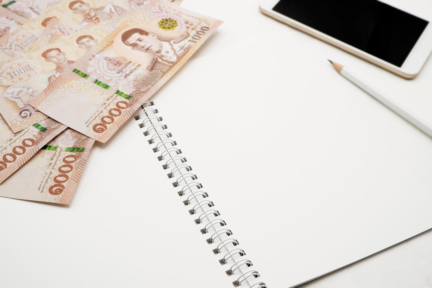 Vide blanc spirale carnet avec blanc crayon, mobile téléphone et pile de Nouveau 1000 thaïlandais baht billets de banque. affaires et la finance concept. photo