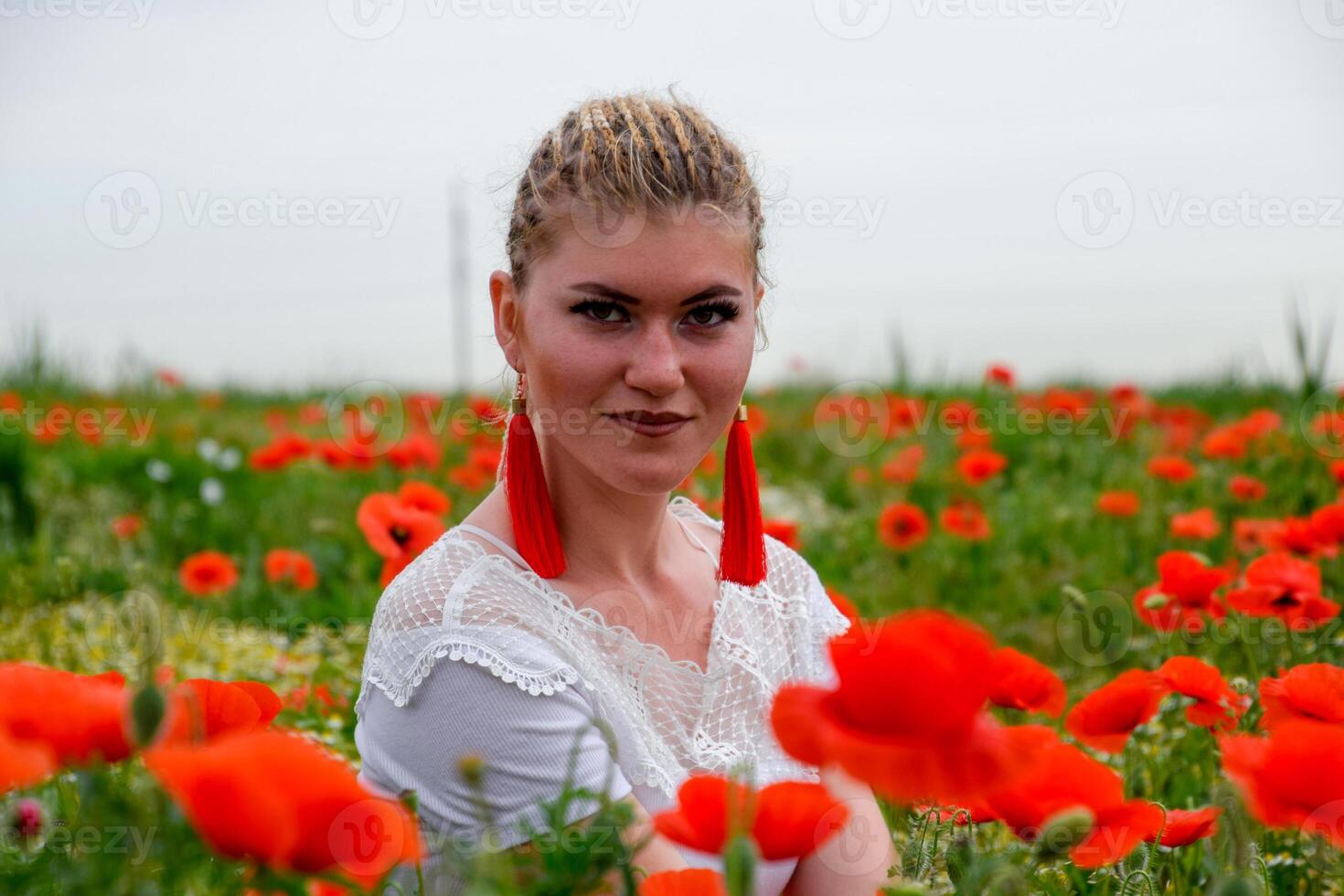 blond Jeune femme dans rouge jupe et blanc chemise, rouge des boucles d'oreilles est dans le milieu de une coquelicot champ. photo