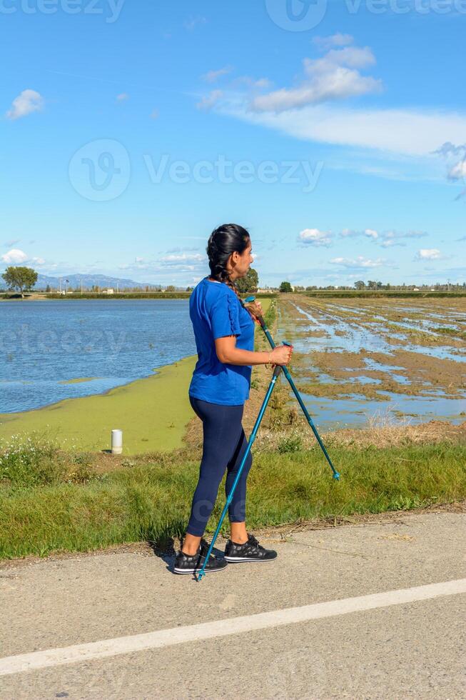 latina femme, actif individuel avec en marchant poteaux suivant à une l'eau corps sur une ensoleillé jour, hispanique latina femme en marchant avec trekking poteaux dans le Èbre delta Naturel parc, Tarragone, catalogne, Espagne photo