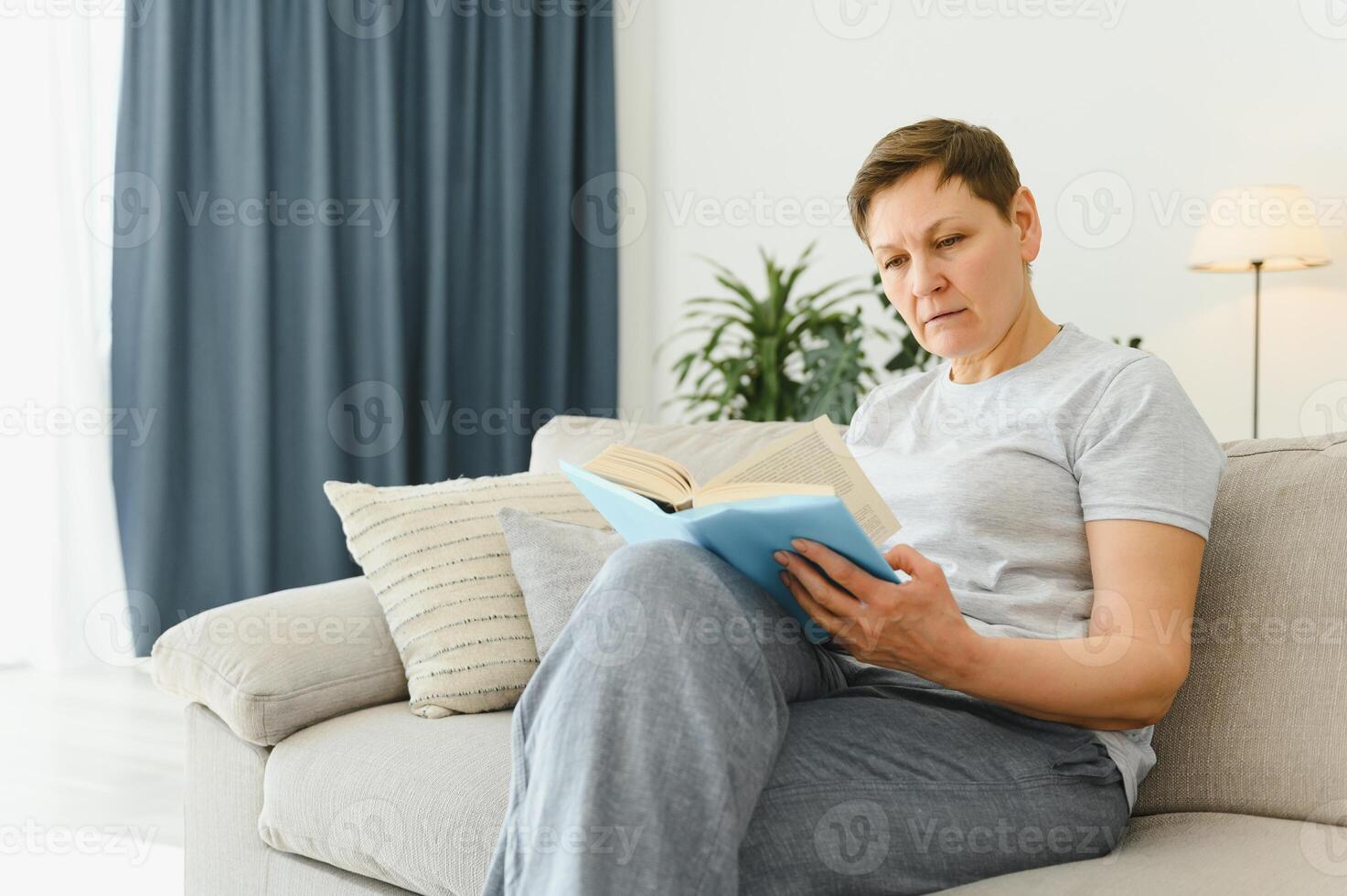 portrait de une content mature femme relaxant sur canapé et en train de lire une livre dans une moderne maison photo