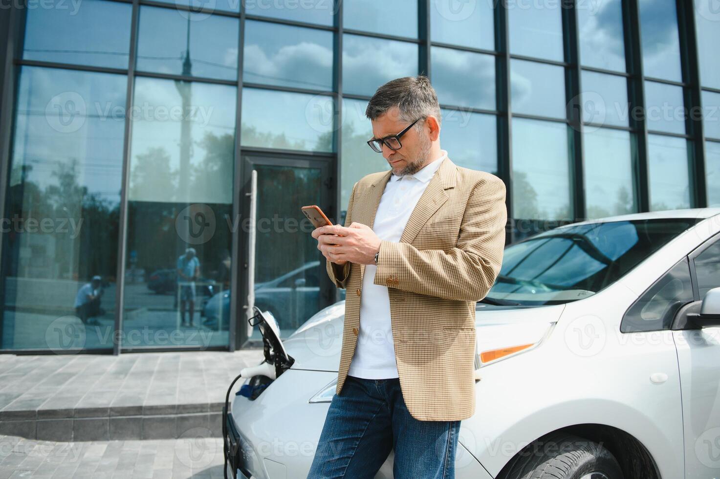 le gars Sam vers le bas sur le capuche de le auto. le sien voiture est mise en charge à le mise en charge gare. une homme regards à le téléphone intelligent écran et sourit. photo