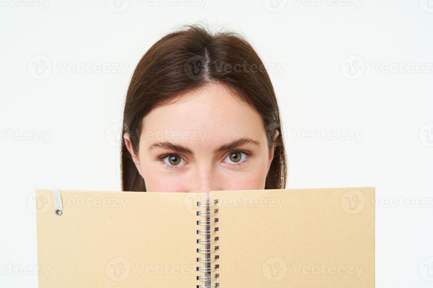 portrait de mignonne Jeune femme cache sa visage derrière planificateur, détient carnet contre sa visage et sourires, isolé plus de blanc Contexte photo