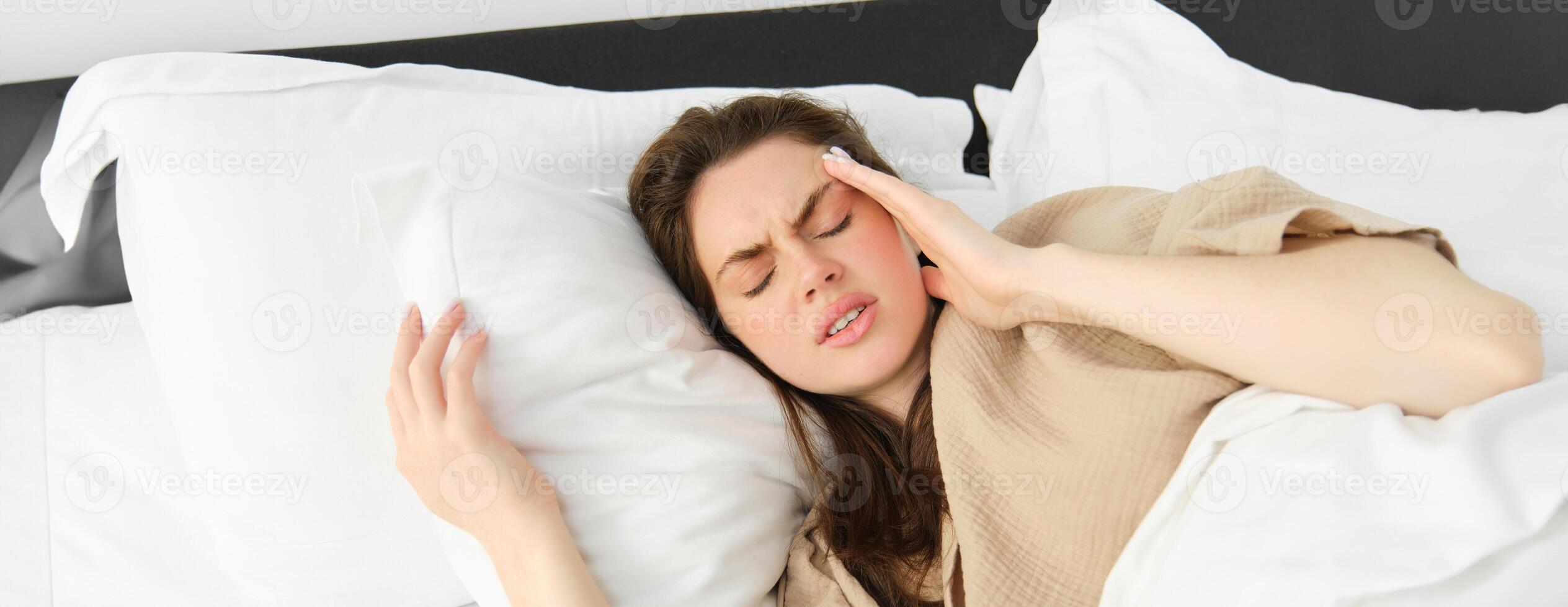 proche en haut portrait de malade femme mensonge dans sa lit et sentiment indisposé, émouvant diriger, a mal de tête, à la recherche frustré de migraine, portant pyjamas photo