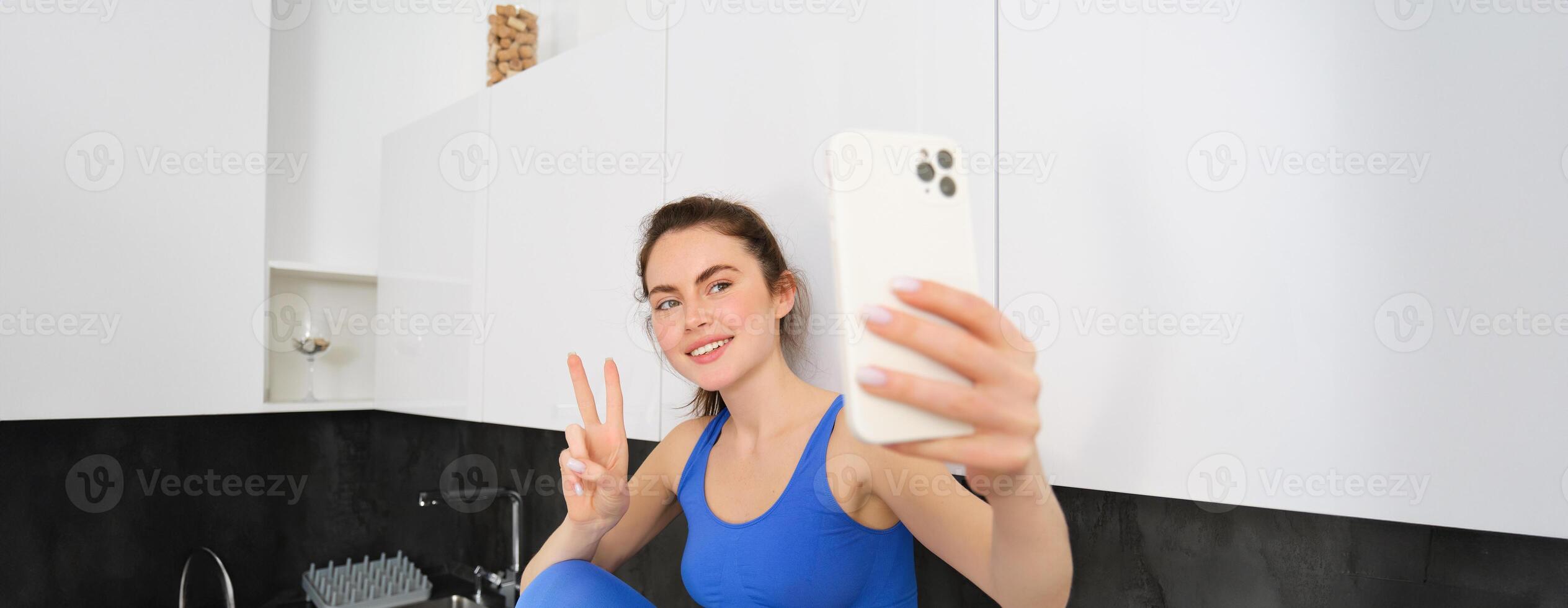 portrait de magnifique femme, aptitude blogueur, prise selfie dans soutien-gorge de sport et leggings, posant dans cuisine avec téléphone intelligent photo