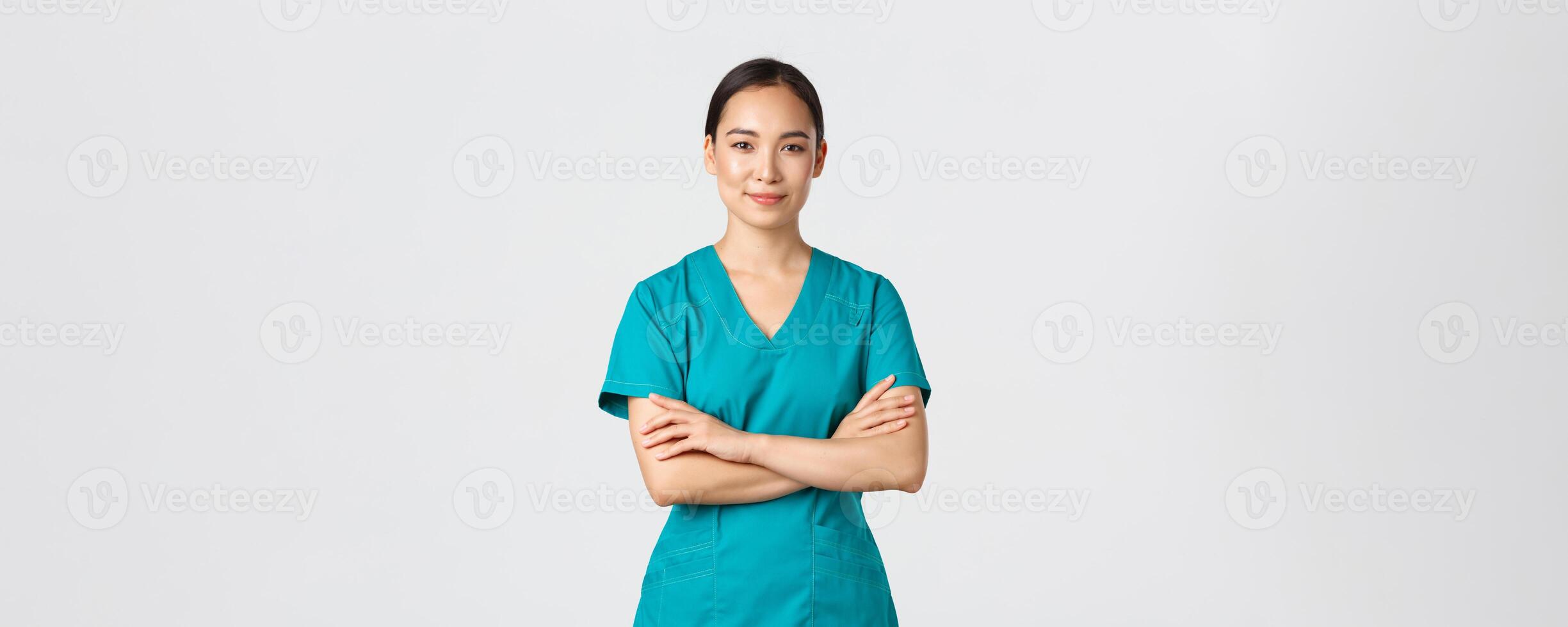 COVID-19 [feminine, soins de santé ouvriers, pandémie concept. sur de soi souriant asiatique infirmière dans gommages permanent sûr de soi, traverser bras poitrine. professionnel médecin prêt pour chirurgie, permanent blanc Contexte photo