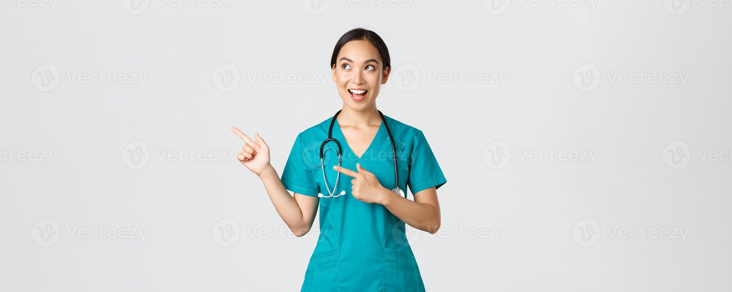 COVID-19 [feminine, soins de santé ouvriers, pandémie concept. optimiste souriant femelle asiatique infirmière, médecin dans gommages, à la recherche et montrer du doigt plus haut la gauche coin, montrant bannière, démontrer annonce photo