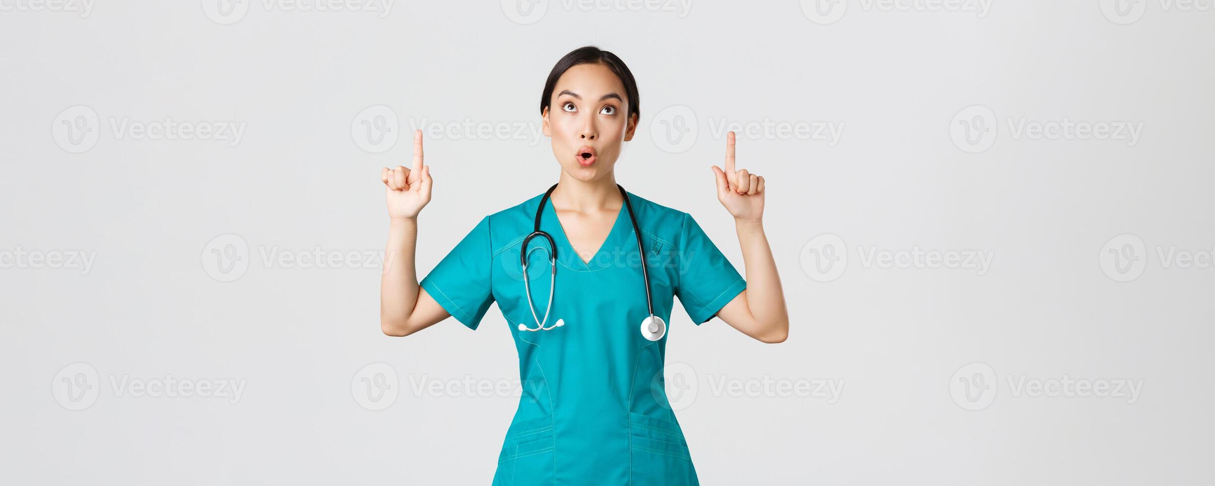 COVID-19 [feminine, soins de santé ouvriers, pandémie concept. impressionné et fasciné jolie asiatique femelle infirmière, médecin dans gommages montrer du doigt et à la recherche en haut avec chuté mâchoire, rester sensationnel étonné photo