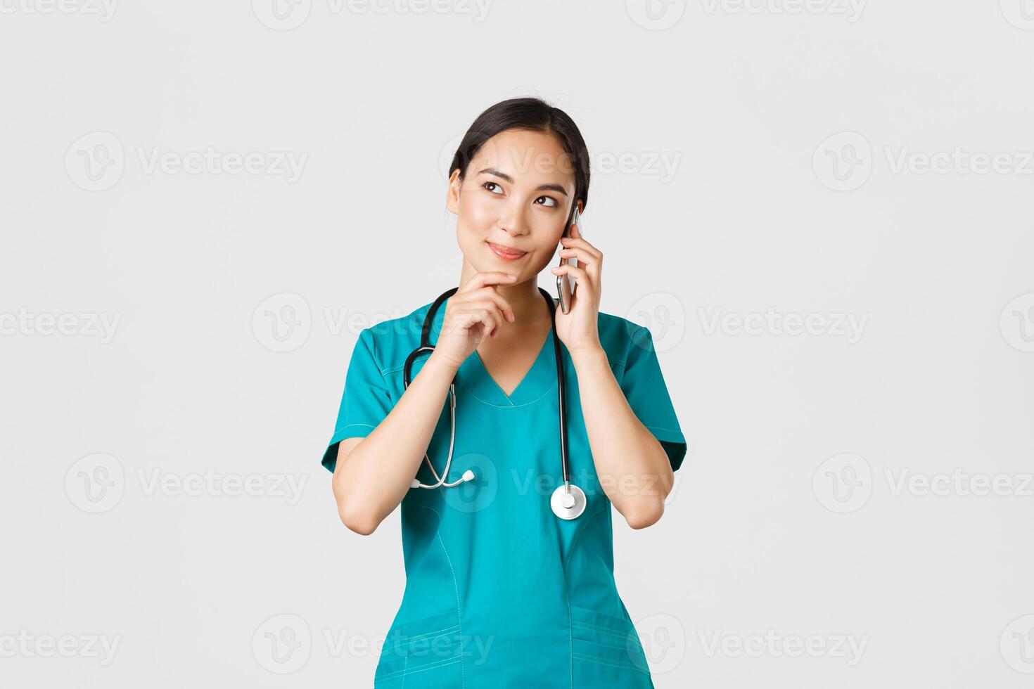 COVID-19 [feminine, soins de santé ouvriers et empêcher virus concept. portrait de souriant asiatique femelle médecin, interne dans gommages parlant sur téléphone et à la recherche réfléchi, en pensant ou fabrication choix photo