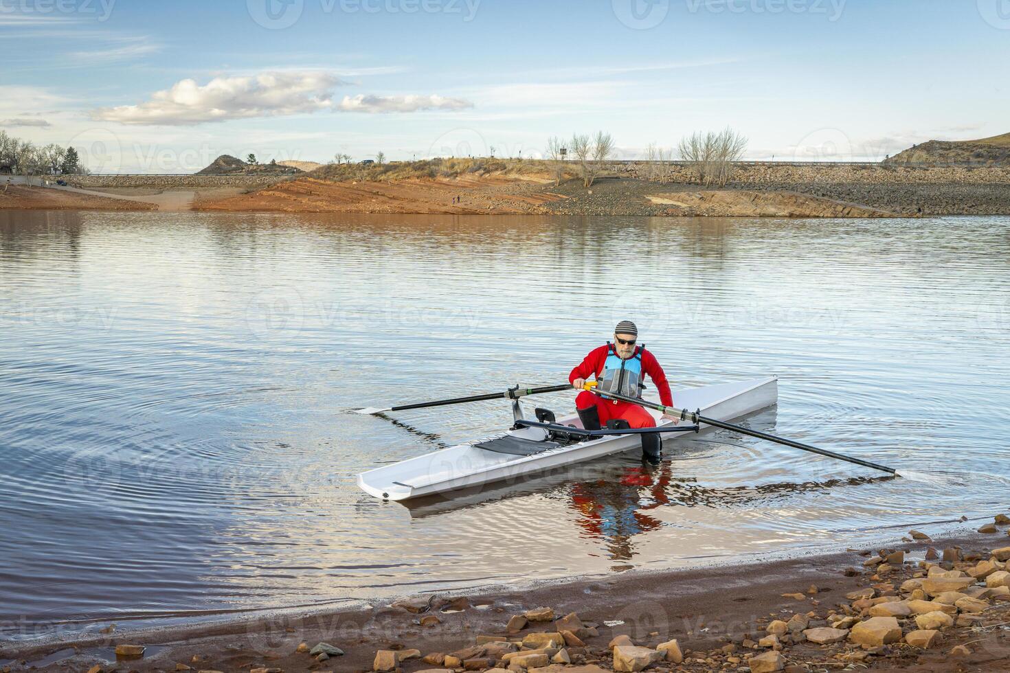 Sénior Masculin rameur dans une côtier aviron coquille est atterrissage sur une rocheux rive de dent de cheval réservoir dans tomber ou hiver paysage dans nord Colorado. photo