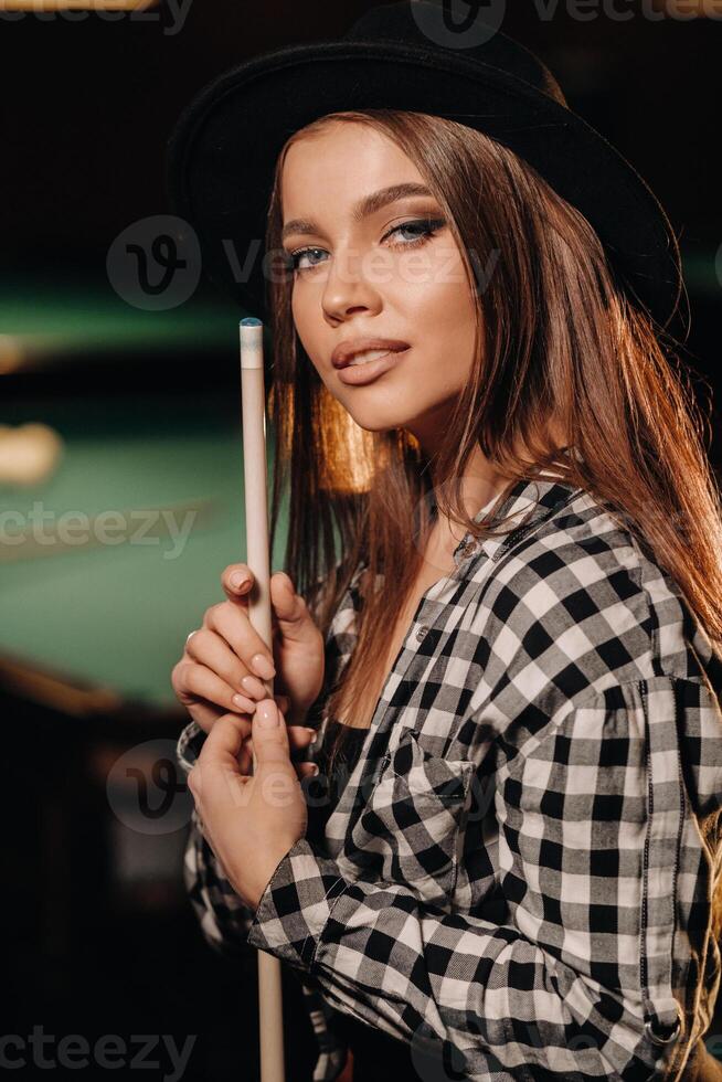 une fille dans une chapeau dans une billard club avec une queue dans sa mains.billard Jeu photo