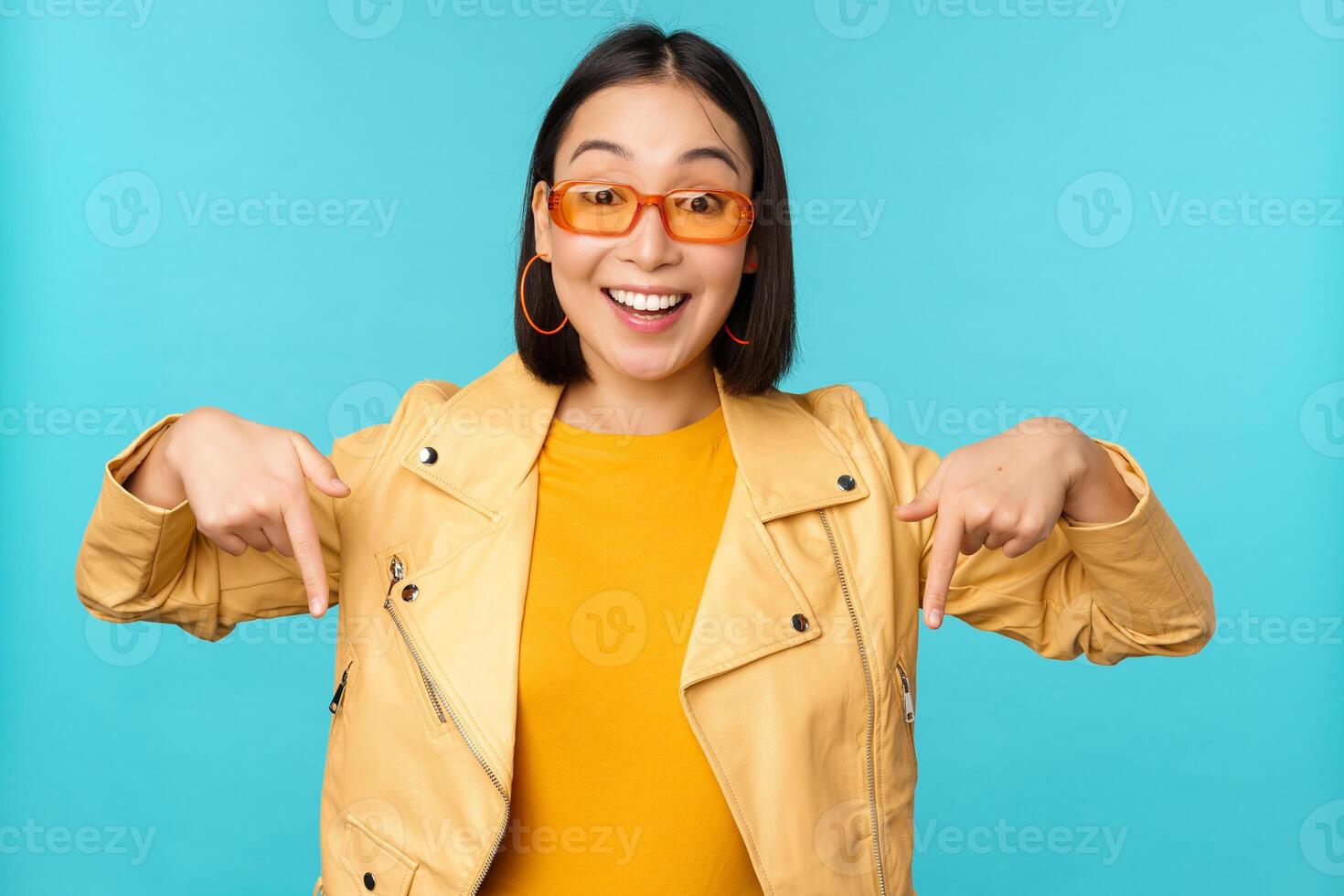 portrait de élégant asiatique fille, porte des lunettes de soleil, sourit et points les doigts bas, spectacles publicité, des stands plus de bleu Contexte photo