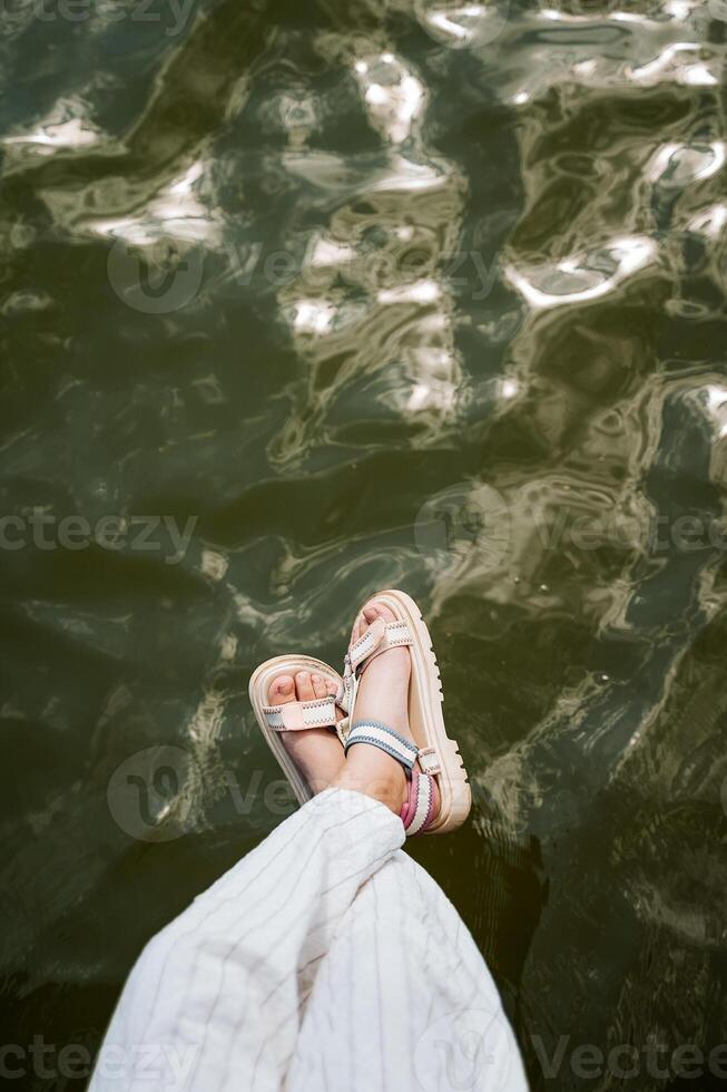 une fille dans lumière rayé un pantalon accroché sa jambes dans des sandales plus de le l'eau surface. photo