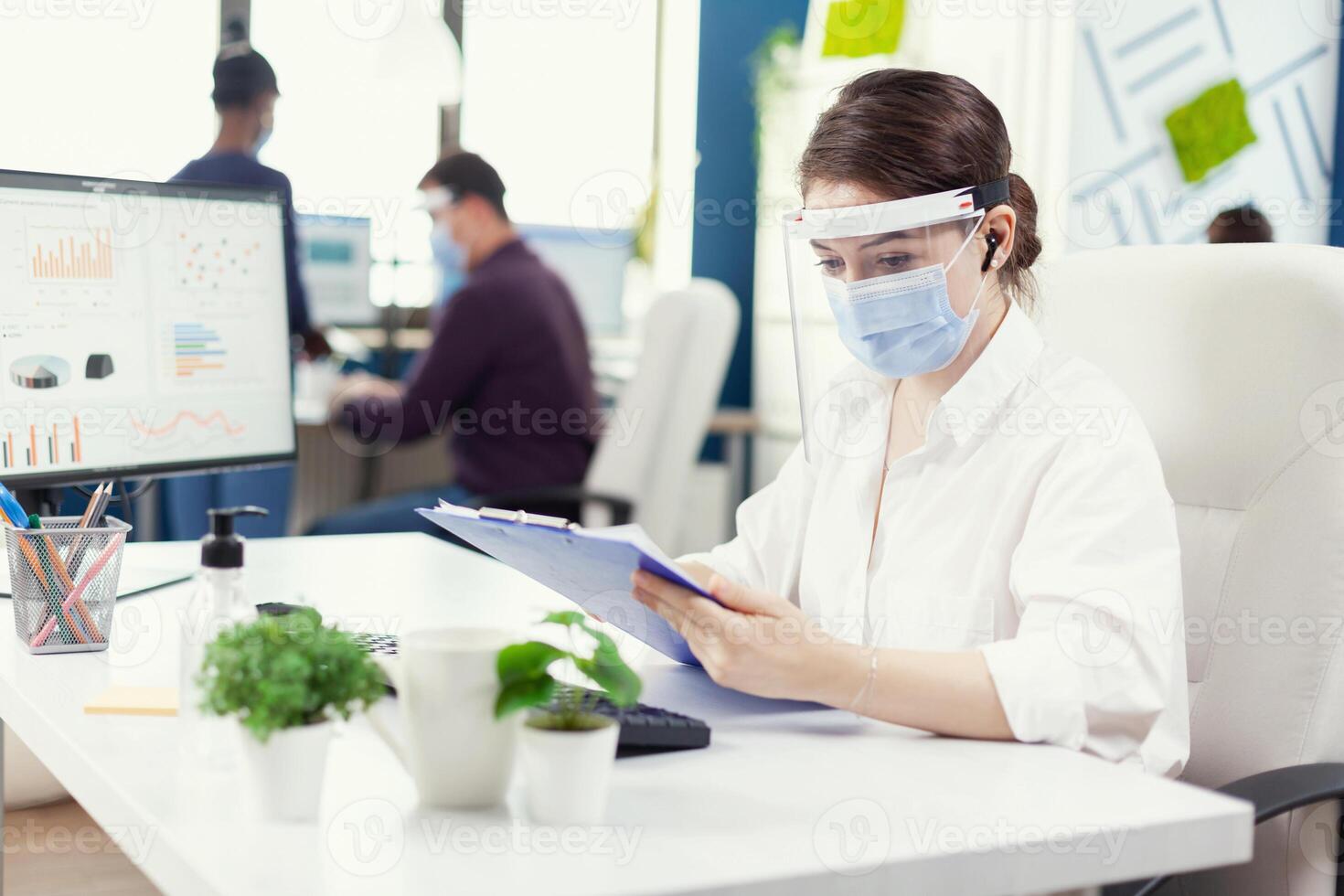 financier conseiller avec sans fil écouteurs vérification financier statistique dans occupé Bureau avec visage masque. des employés avec visières travail dans entreprise entreprise espace de travail respectant social distance en cours d'analyse Les données et graphiques. photo