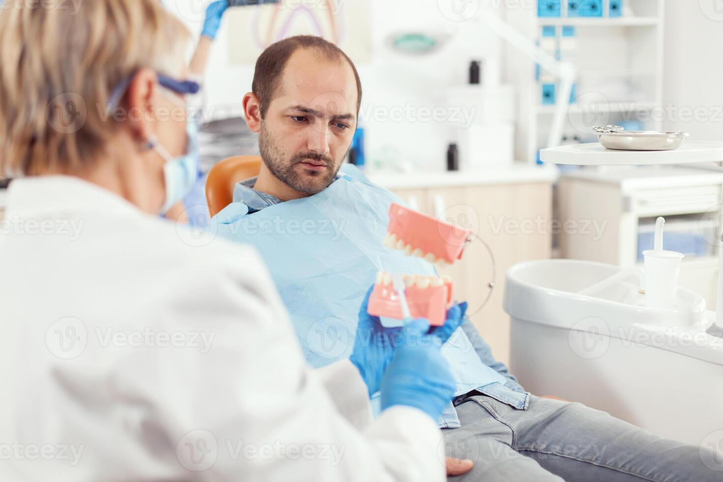 stomatologue expliquant correct dentaire hygiène en utilisant les dents squelette pendant stomatologie rendez-vous. dentiste en train de préparer malade homme patient pour dentisterie chirurgie séance sur chaise dans examen pièce photo