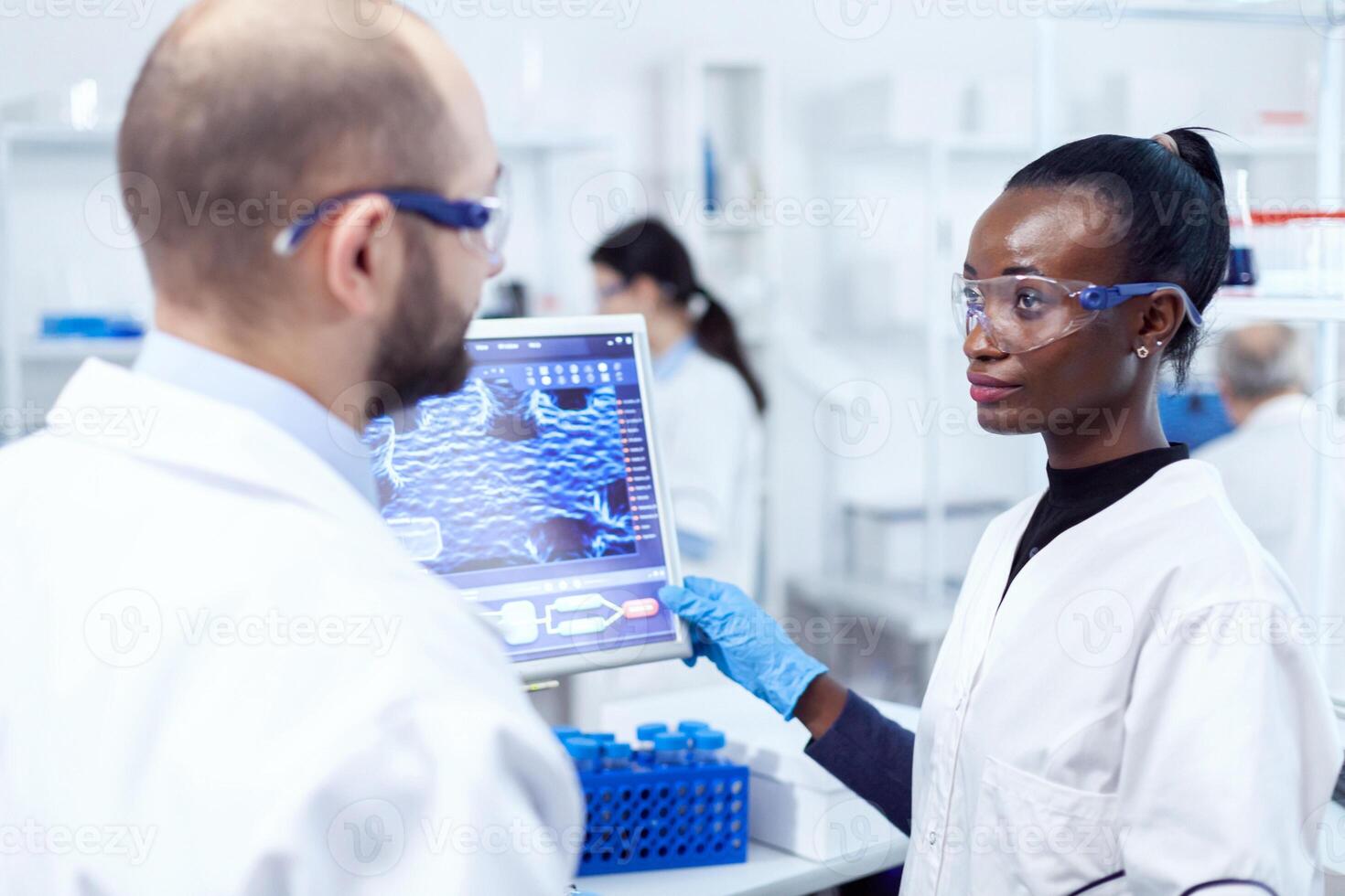 africain scientifique discuter résultat de virus recherche avec superviseur dans laboratoire en utilisant ordinateur. Multi-éthnique équipe de médical des chercheurs travail ensemble dans stérile laboratoire portant protection des lunettes et gants. photo