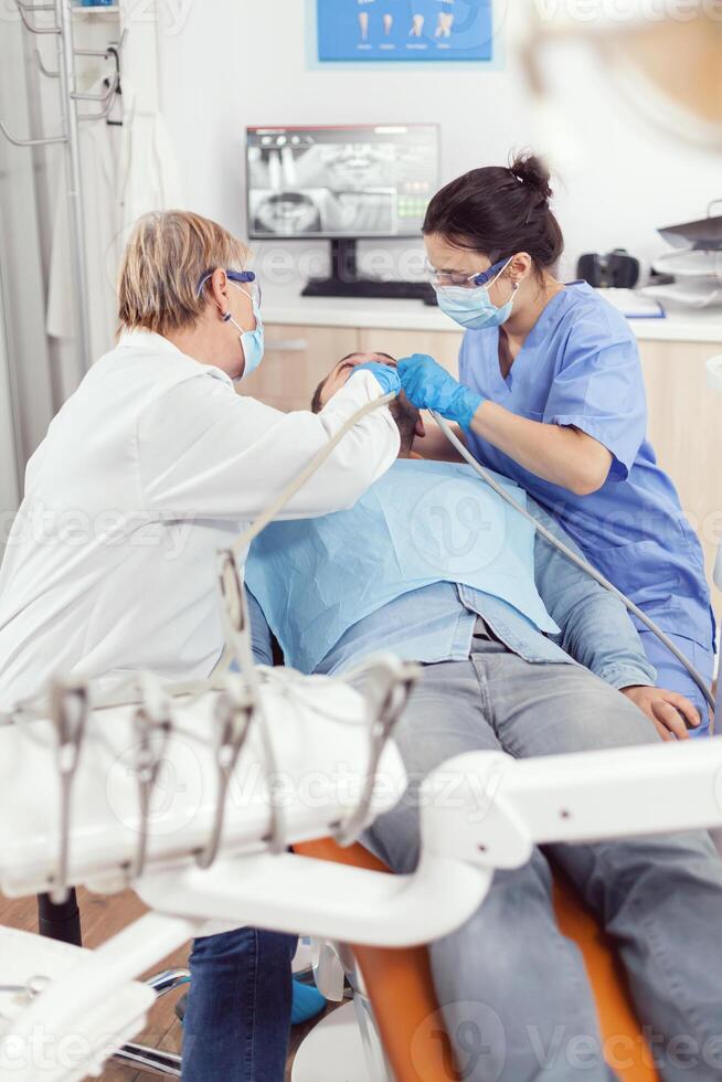 orthodontiste Sénior femme Faire stomatologie traitement sur malade patient en train de préparer pour dent chirurgie. homme implantation sur dentaire chaise dans hôpital orthodontique Bureau pendant médical procédure photo