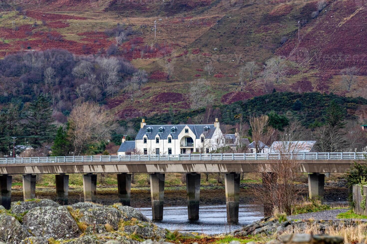 pittoresque blanc maison avec une ardoise toit derrière une pierre pont plus de une tranquille rivière, ensemble contre une toile de fond de roulant collines et clairsemé des bois dans Écosse. photo