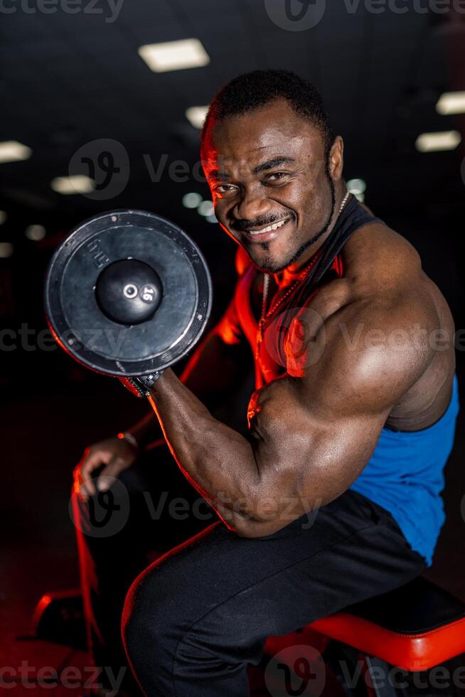 fort souriant bodybuilder dans bleu T-shirt exercice avec lourd haltère dans moderne foncé salle de sport. photo