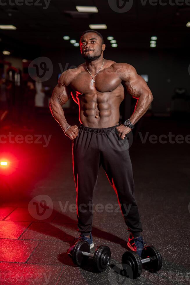 à moitié nu fort africain américain bodybuilder permanent avec sport équipement dans foncé moderne salle de sport. photo