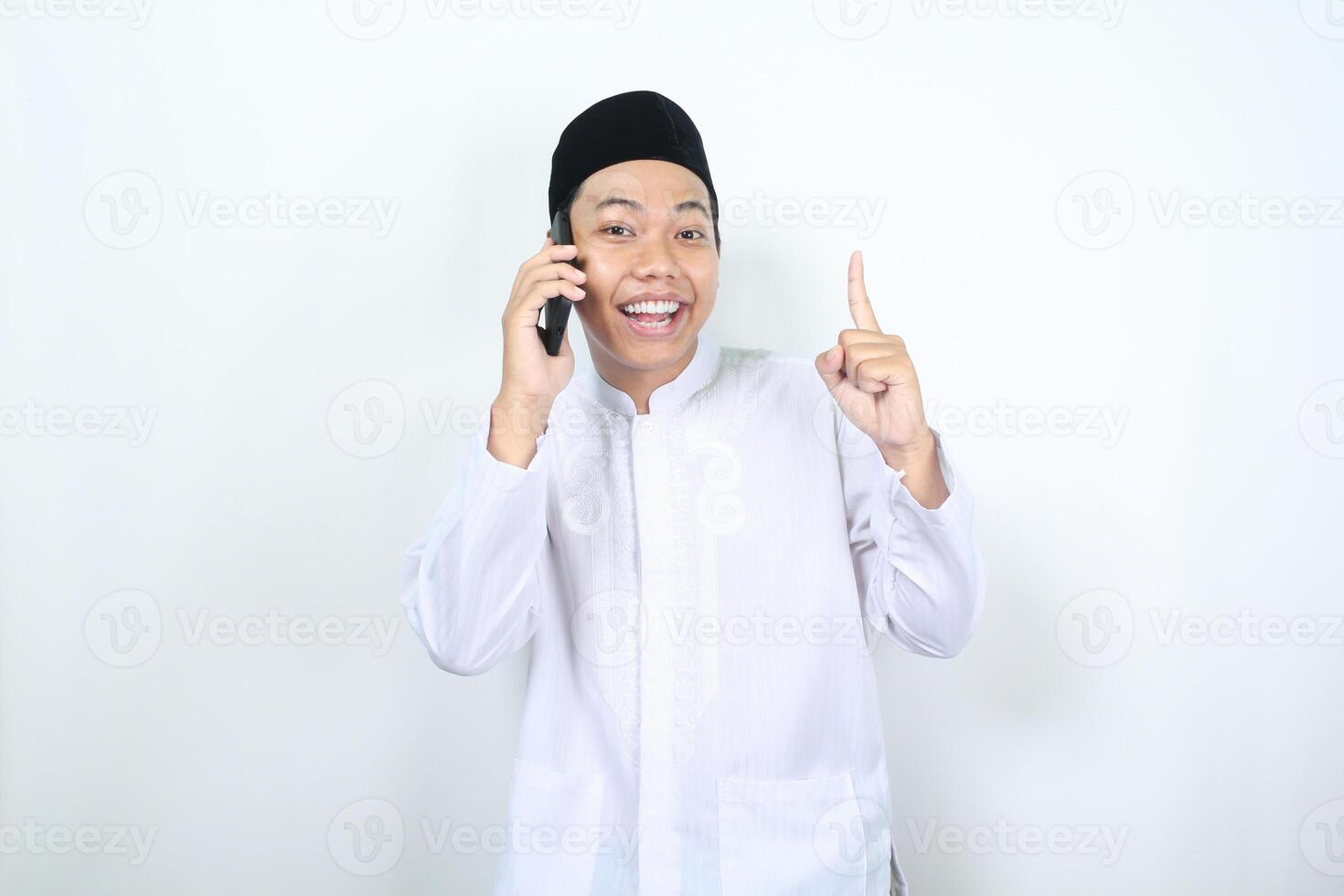 de bonne humeur musulman asiatique homme montrer du doigt à au dessus tandis que Parlant sur le sien téléphone avec souriant visage isolé photo