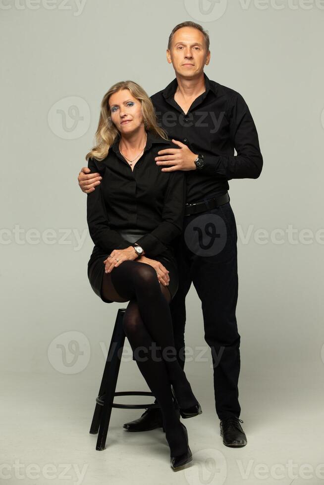 portrait de une mature couple. couple, deux copains un personnes âgées aux cheveux gris homme et une blond femme dans noir vêtements poser, isolé sur une gris arrière-plan, studio portrait photo