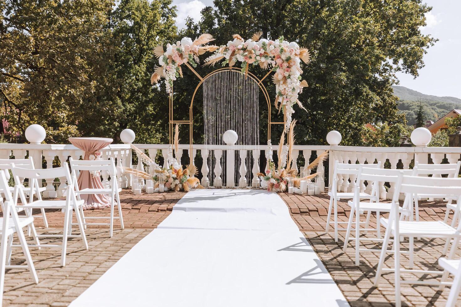 d'or cambre décoré avec fleurs sur le Contexte de des arbres. une blanc chemin cette pistes à le cambre, beaucoup blanc chaises. préparation pour le mariage la cérémonie photo