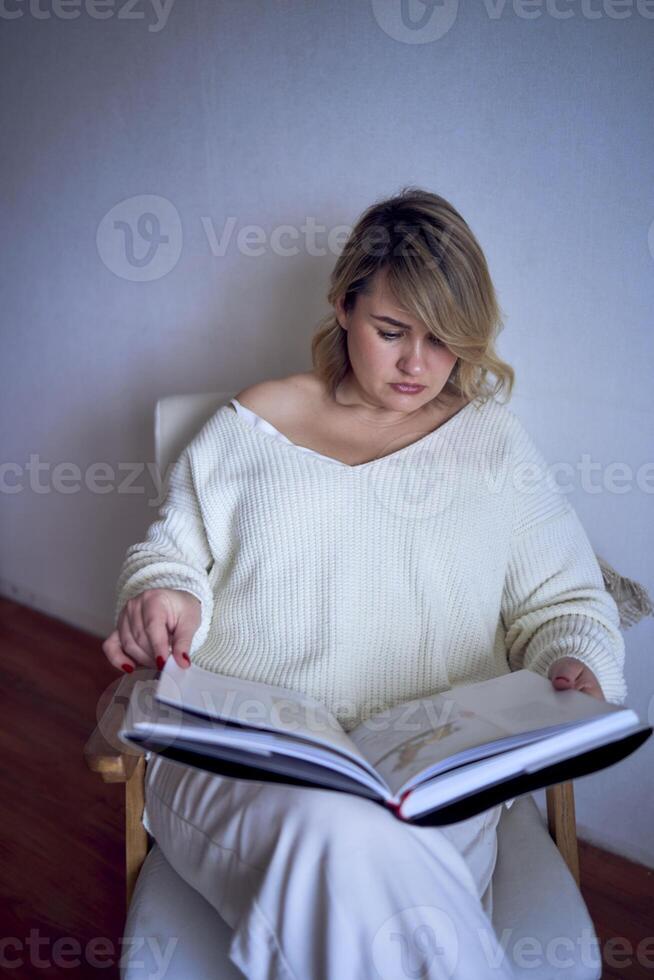 une de taille moyenne femme dans lumière vêtements lit une livre tandis que séance dans une blanc chaise dans une lumière pièce photo