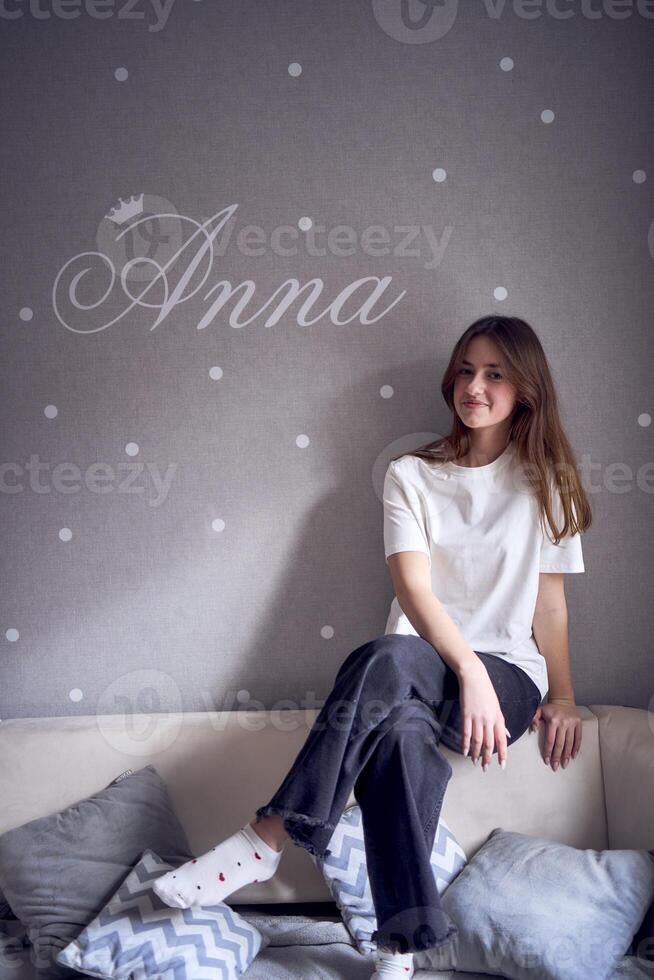 une magnifique adolescent fille est assis sur une lit près le mur avec sa Nom écrit sur il photo
