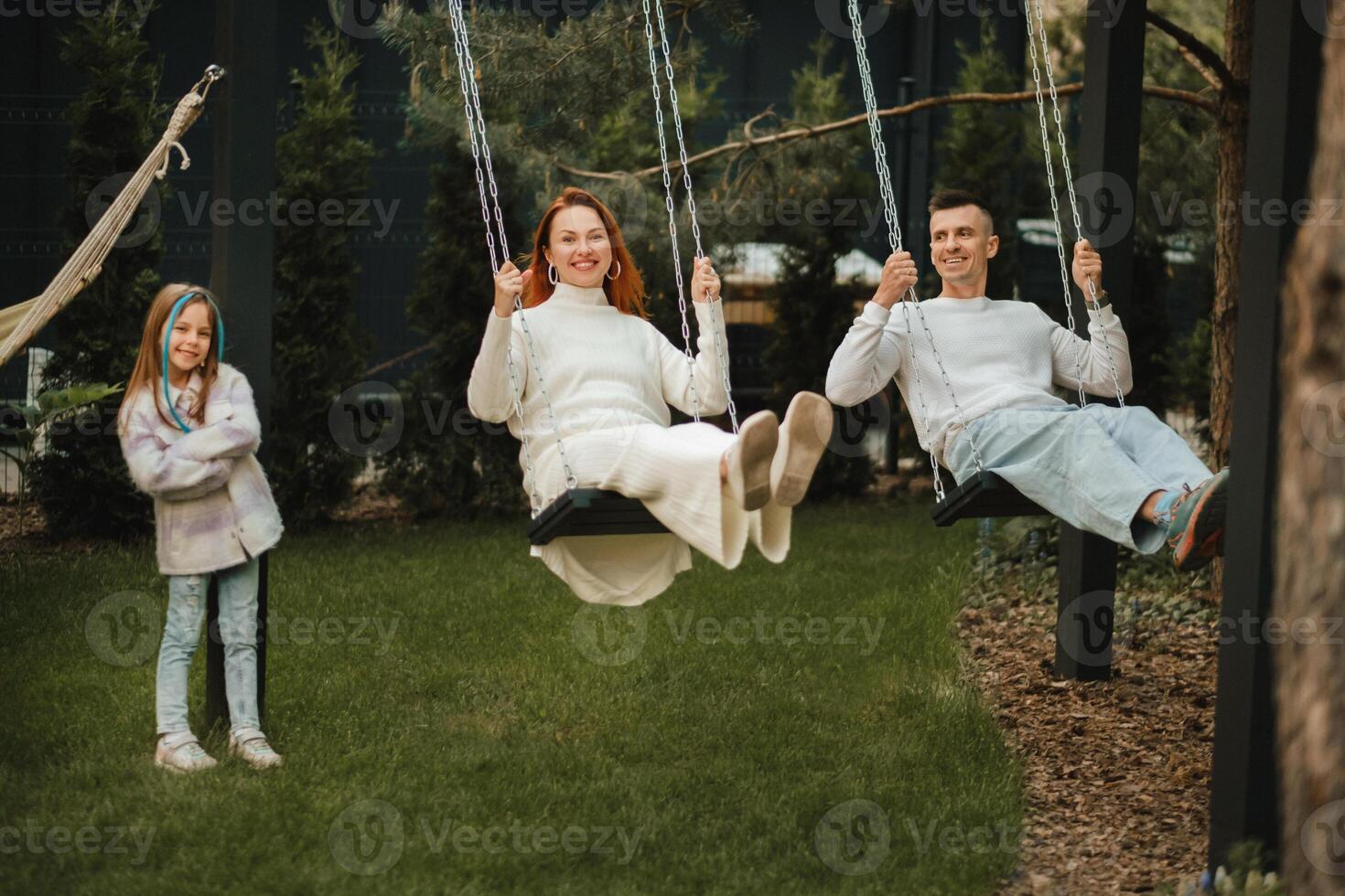 maman et papa sont équitation sur une balançoire et là est une fille permanent suivant à eux. le famille est repos sur une balançoire photo