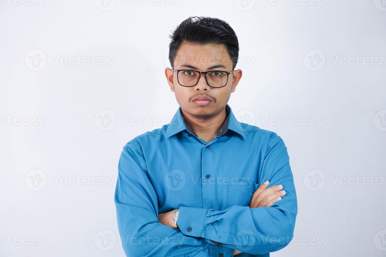 en colère asiatique homme avec des lunettes permanent avec franchi bras et à la recherche à caméra portant bleu chemise isolé sur blanc Contexte photo