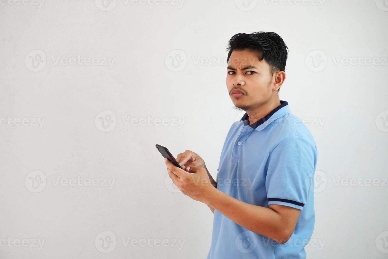 Jeune asiatique homme permanent en portant une téléphone avec un en colère et désapprobateur expression portant bleu t chemise isolé sur blanc Contexte photo