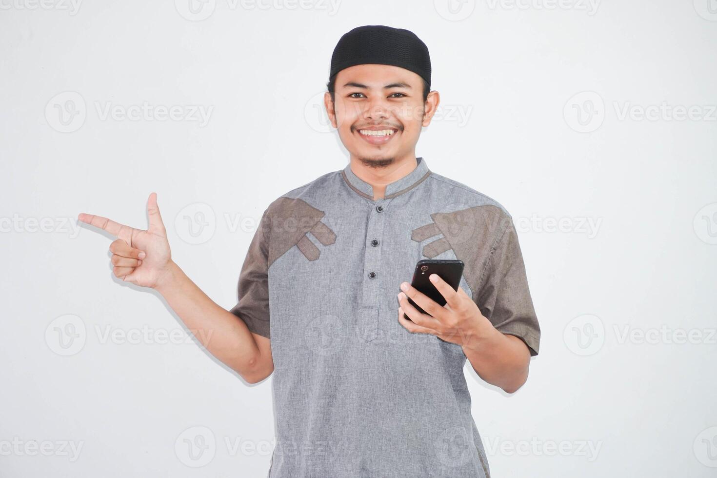 souriant ou content asiatique musulman homme en portant mobile téléphone avec doigt montrer du doigt côté portant musulman vêtements isolé sur blanc Contexte photo