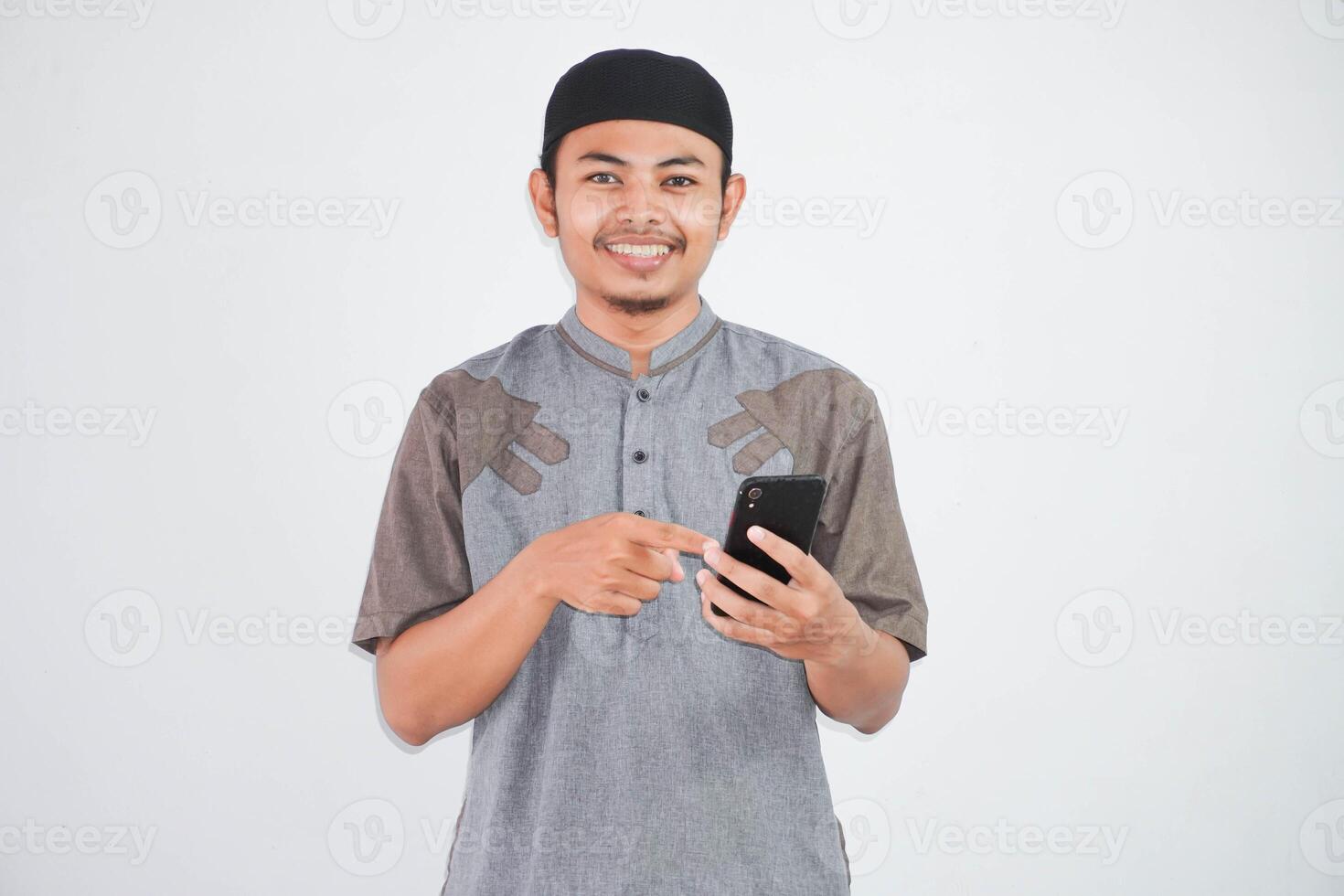 souriant asiatique musulman homme en portant et doigt montrer du doigt mobile téléphone portant gris musulman vêtements isolé sur blanc Contexte photo