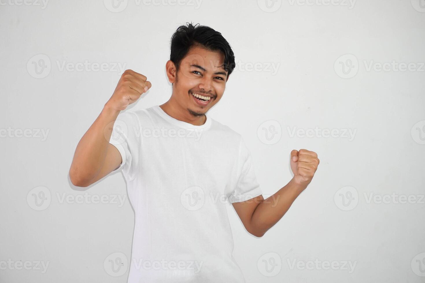 sur de soi de bonne humeur Beau asiatique homme levage mains en haut portant blanc t chemise gagnant geste serrant poings. se sent bonheur spectacle poing en haut Succès isolé sur blanc Contexte photo