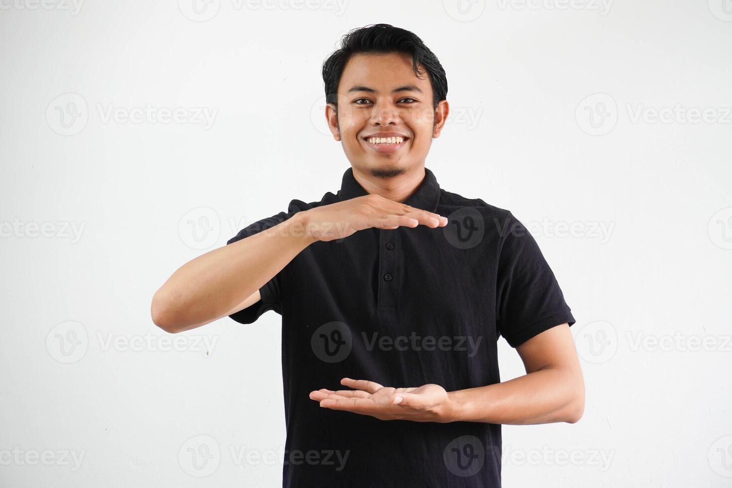 Jeune asiatique homme contre une blanc studio Contexte portant noir polo t chemise, en portant quelque chose avec tous les deux mains, produit présentation. photo