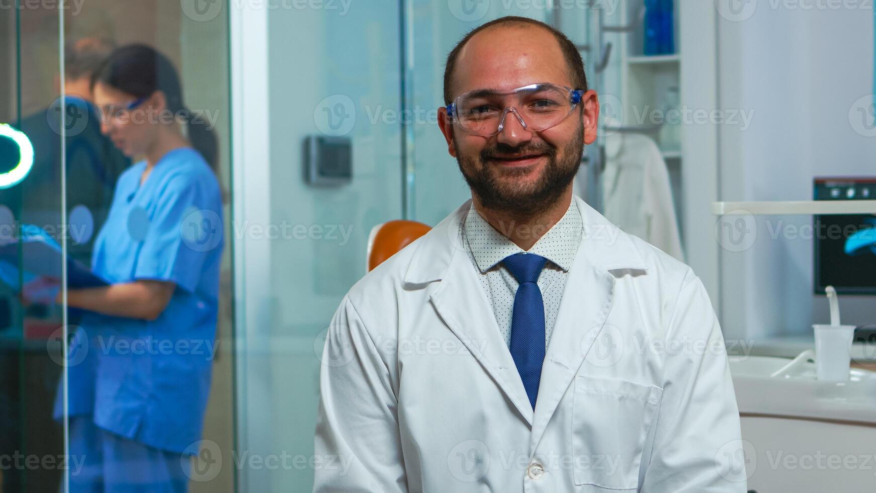 portrait de stomatologue souriant à caméra étant dans dentaire Bureau tandis que infirmière est parlant avec patient dans Contexte. dentisterie médecin à la recherche sur webcam séance sur chaise dans stomatologique clinique. photo