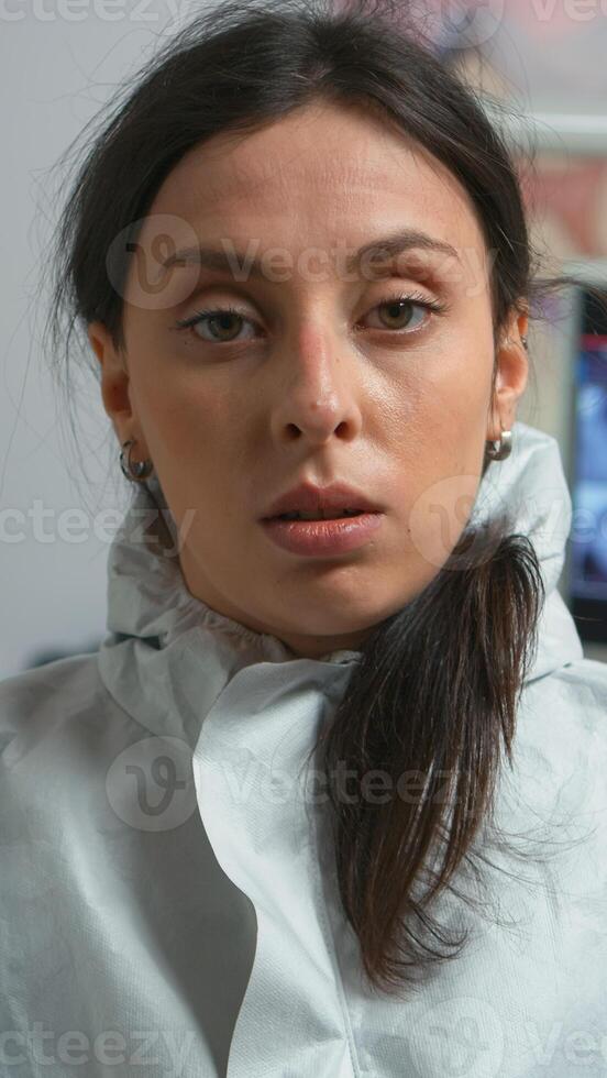 brut précaire portret de fatigué médecin dans combinaison prise de le masque à la recherche sur caméra après examiner patient pendant coronavirus pandémie. dentiste infirmière portant combinaison, visage bouclier, masque, gants dans stomatologique bureau. photo